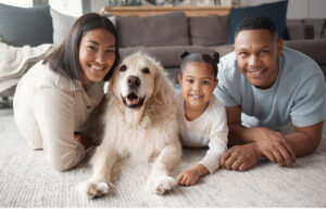 Portrait of a happy mixed race family of three relaxing on the lounge floor with their dog. Loving black family being affectionate with a foster animal. Young couple bonding with their daughter and rescued puppy at home